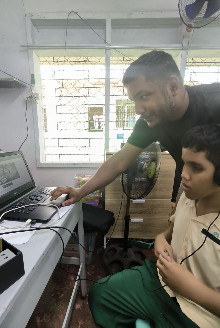 A teacher operates the laptop next to a student listening through headphones.